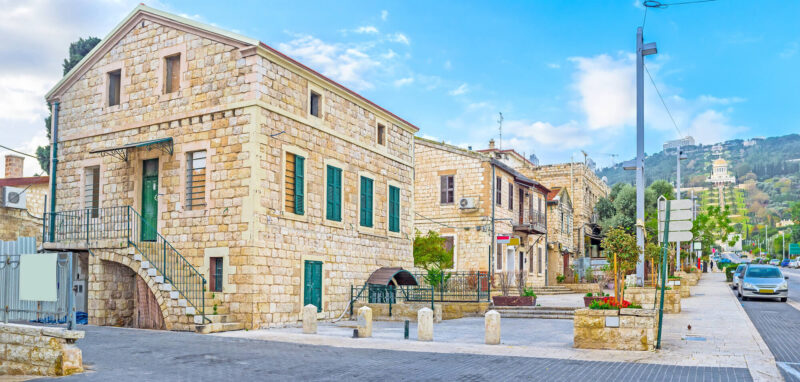 תמונת נושא של המאמר דירה להשכרה בחיפה המציגה רחוב מרוצף אבנים באזור היסטורי של חיפה עם בנייני אבן ישנים ובאופק גני הבאהיים.