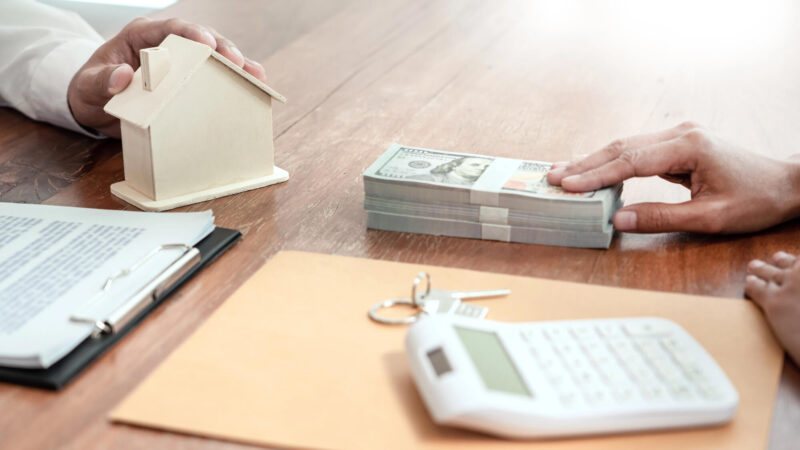 תמונת נושא של המאמר איך להיות יזם נדלן? המציגה על שולחן עץ דגם בית קטן ערימת שטרות דולר ידיים של אנשים מסמכים מחשבון ומפתחות המייצגים עסקת נדל